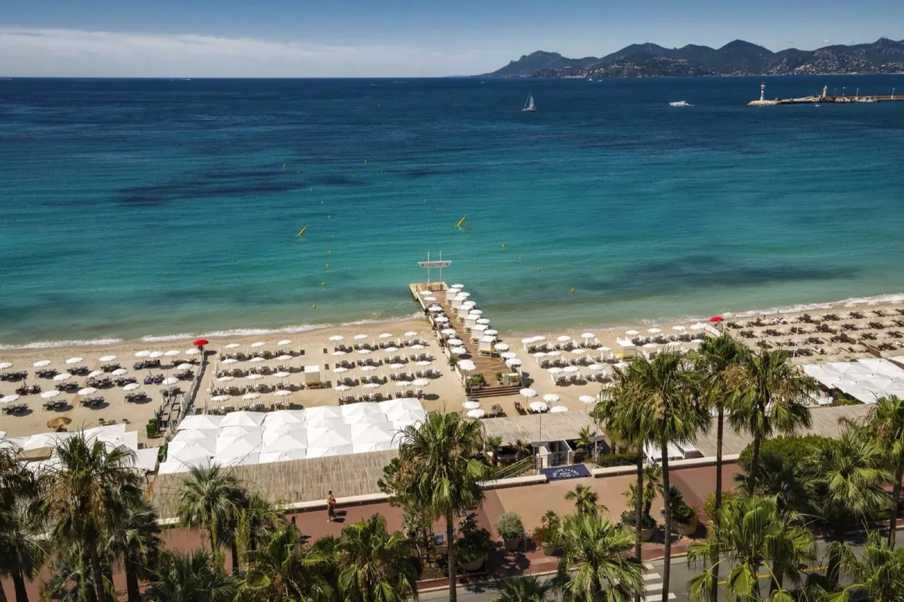 Aerial view of a palm-lined Riviera beach club with rows of white parasols