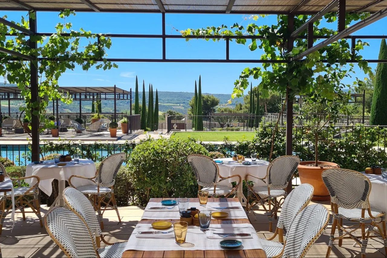 Table set for lunch on a vine-covered terrace overlooking a pool and countryside